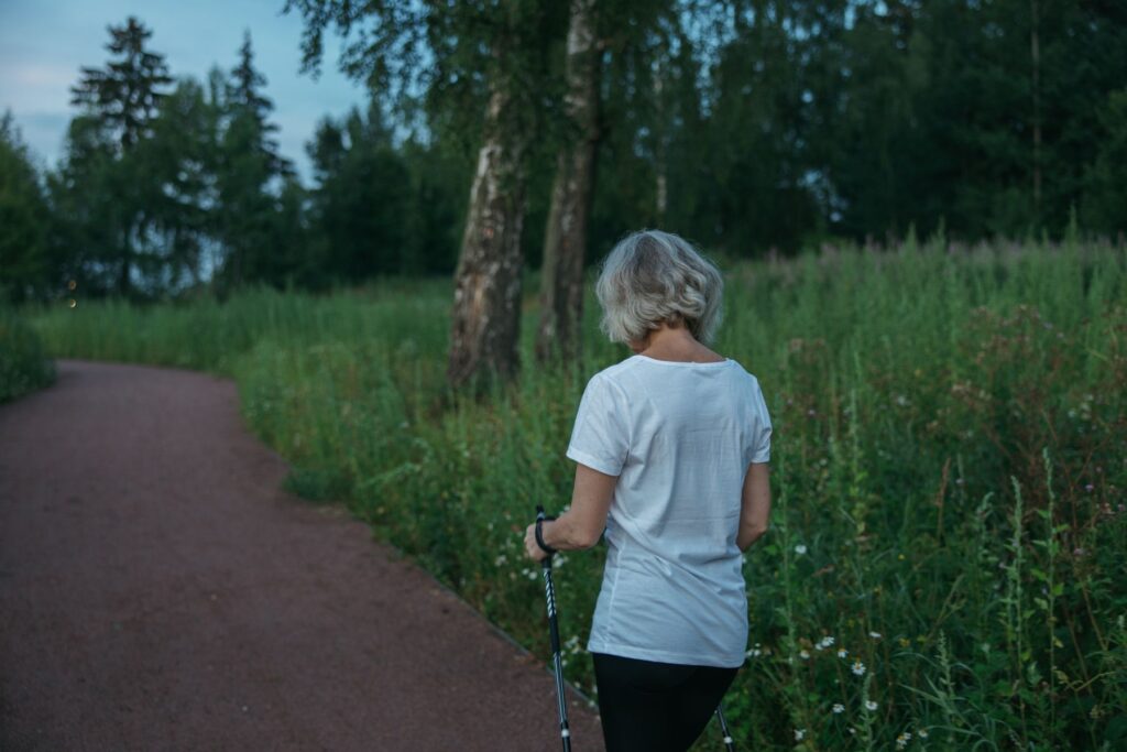 Frau geht mit Nordic-Walking-Stöcken auf einem Parkweg entlang einer grünen Wiese