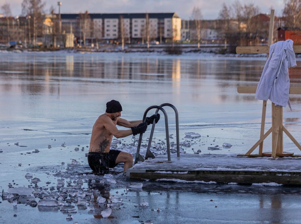 Mann steigt über eine Leiter aus einem Eisloch in einem zugefrorenen See, Eisschollen im Wasser, Bademantel am Ufer