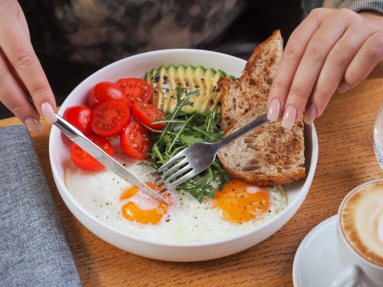 Proteinreiches Frühstück in einer Schüssel: zwei Spiegeleier, Avocado, Tomaten, Rucola und Vollkornbrot, daneben ein Cappuccino