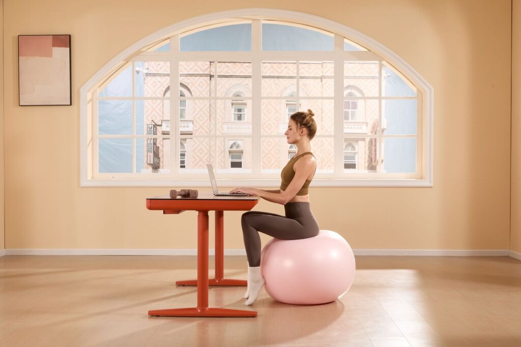 Frau sitzt auf einem Gymnastikball an einem Schreibtisch und arbeitet am Laptop