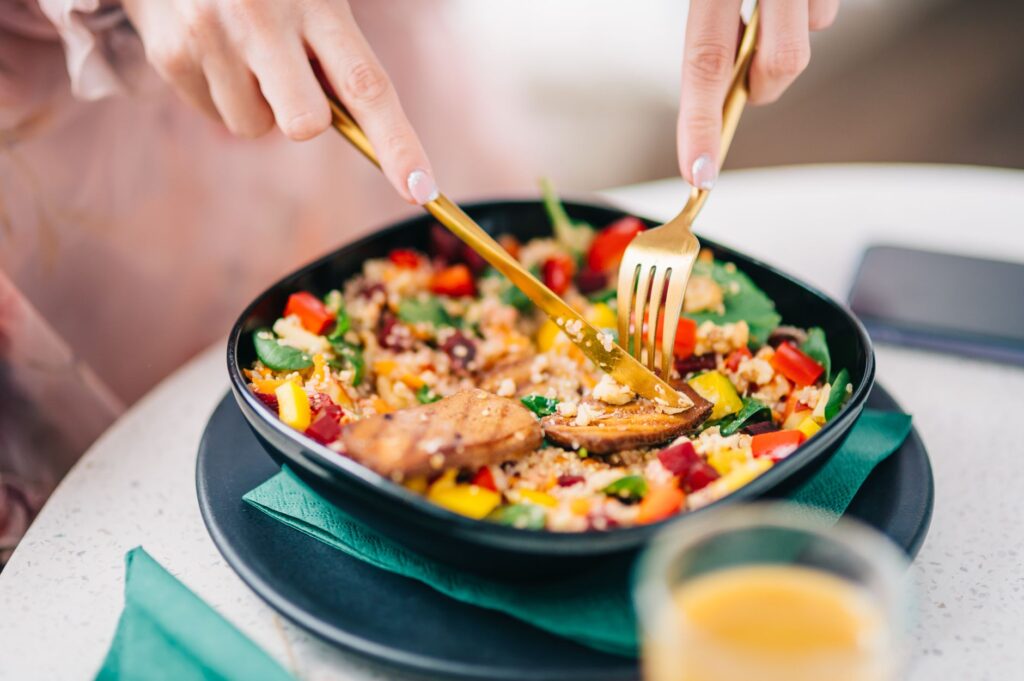 Hände schneiden mit Messer und Gabel eine bunte Quinoa-Bowl mit Gemüse an – Sinnbild für bewusstes, langsames Essen