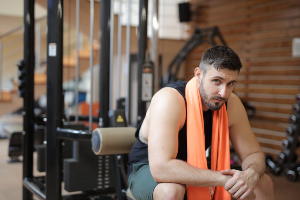 Mann sitzt nach dem Krafttraining mit orangem Handtuch im Fitnessstudio und macht Pause