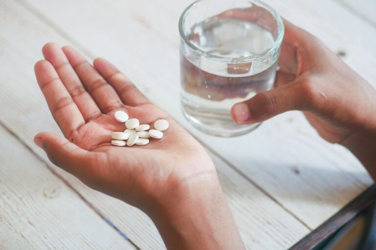 Offene Handfläche mit weißen Tabletten, daneben ein Glas Wasser auf hellem Holztisch