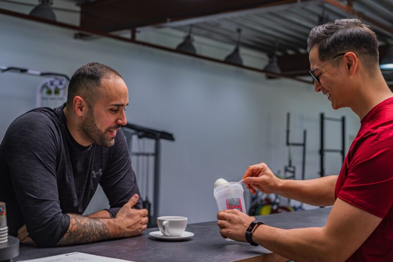 Zwei Männer in einem Fitnessstudio, einer rührt Proteinpulver in einen Shaker