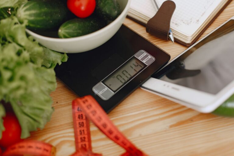 Küchenwaage mit einer Schüssel Gemüse, Notizbuch, Tablet und Maßband auf einem Holztisch