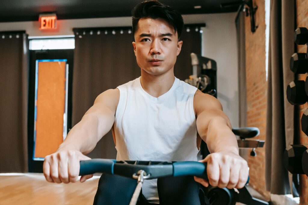 Mann trainiert am Rudergerät im Fitnessstudio, im Hintergrund ein Hantelstaender