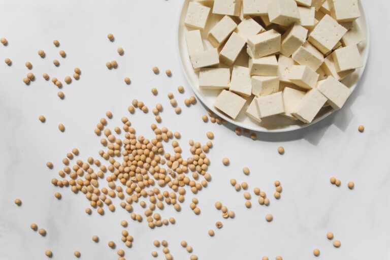 Würfel aus Tofu auf einem weißen Teller neben verstreuten getrockneten Sojabohnen auf heller Marmorfläche