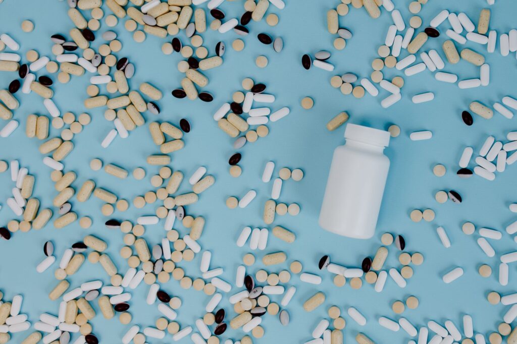 Viele weiße und beige Kapseln und Tabletten rund um eine offene weiße Supplement-Dose auf hellblauem Hintergrund